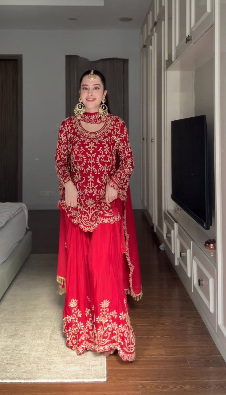 A mannequin displaying a full red embroidered salwar suit with a flowing sharara and a dupatta draped over the shoulder in a boutique setting.