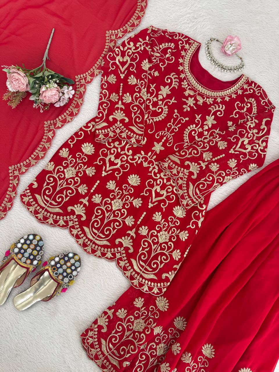 A woman modeling a red pakistani suit dress featuring heavy gold embroidery on the neck and sleeves, paired with large gold earrings and a maang tikka.
